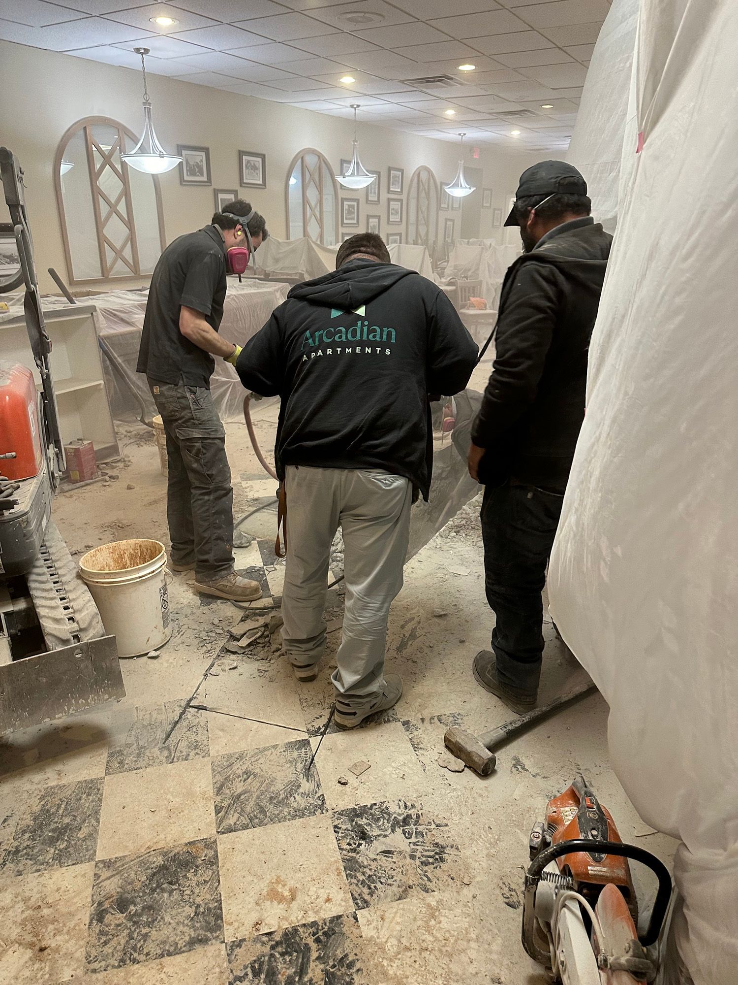 Three construction workers in a dusty interior: one uses a tool, another observes, and the third oversees.