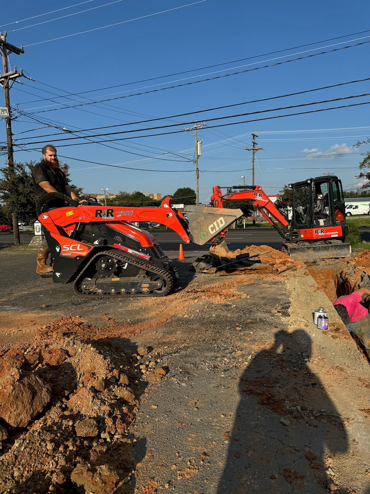 Man operating red skid steer, construction site. Two excavators, dirt, blue sky.