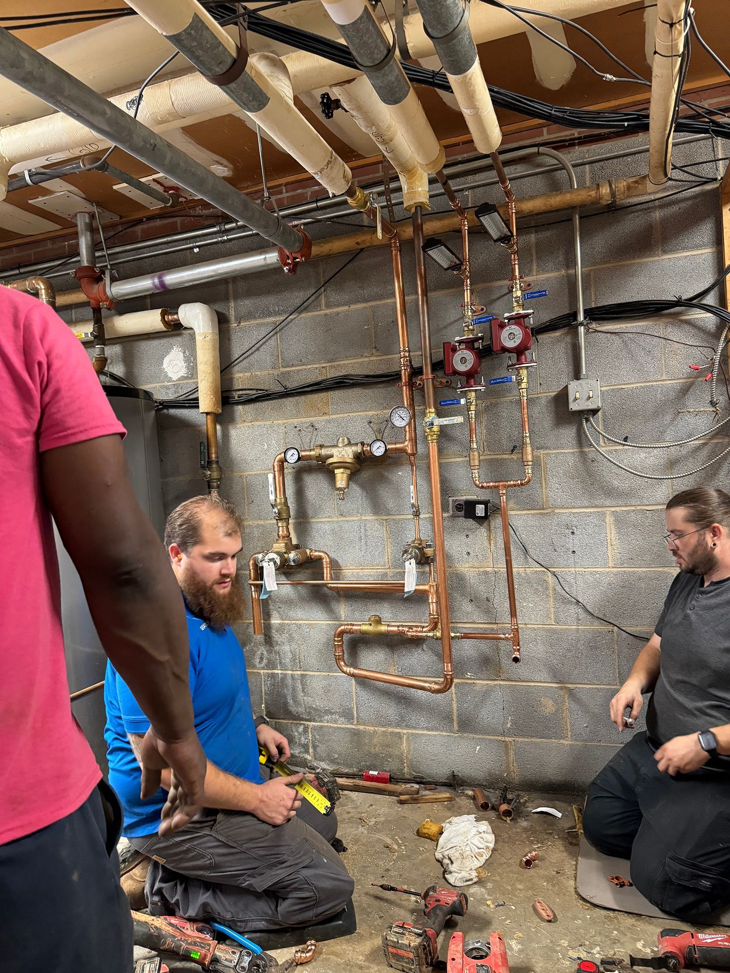 Three plumbers working on copper pipes in a basement, one is kneeling, others are standing.