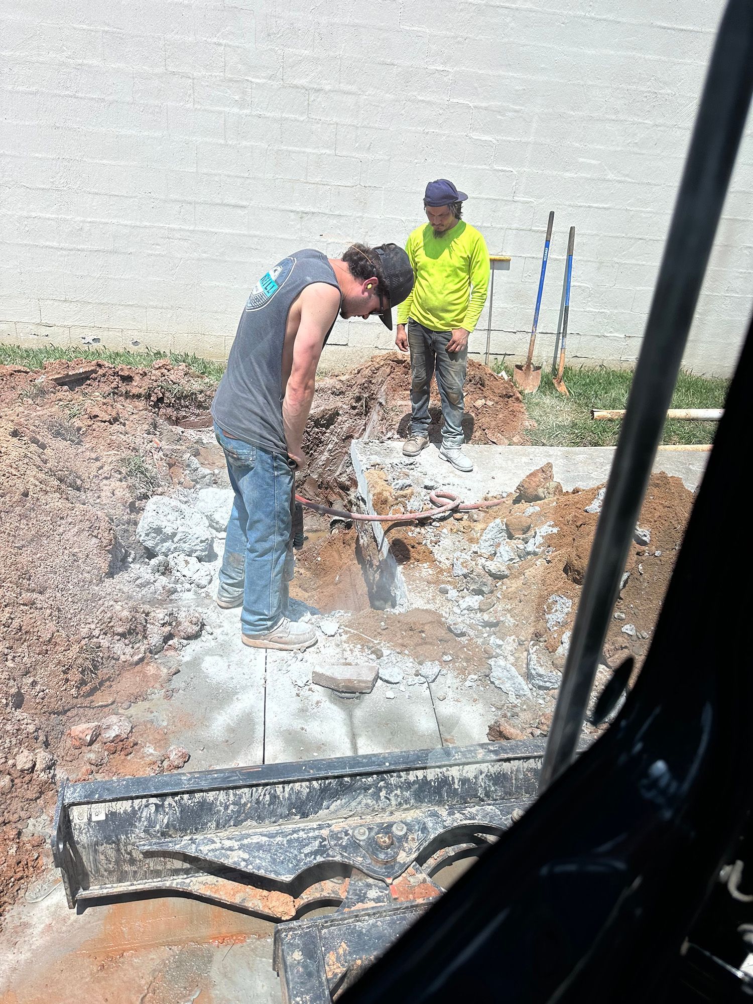 Two construction workers excavate near a white building. One operates a machine, the other looks on.