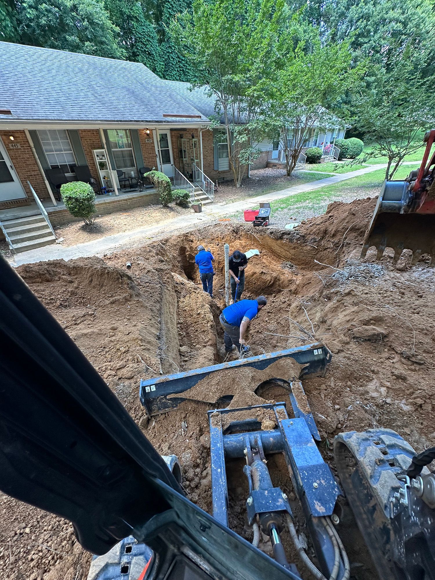 Construction workers in trench, laying pipes near a house, viewed from excavator's cab.