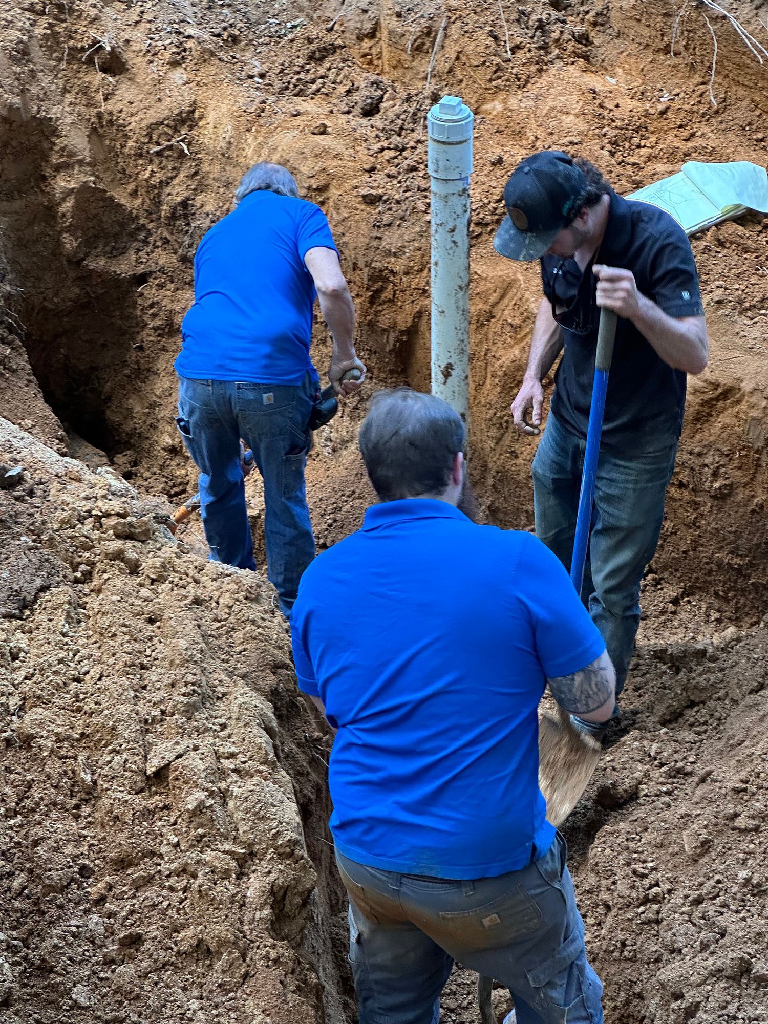 Three people digging in a trench around a vertical pipe. They are wearing blue shirts and a black cap.