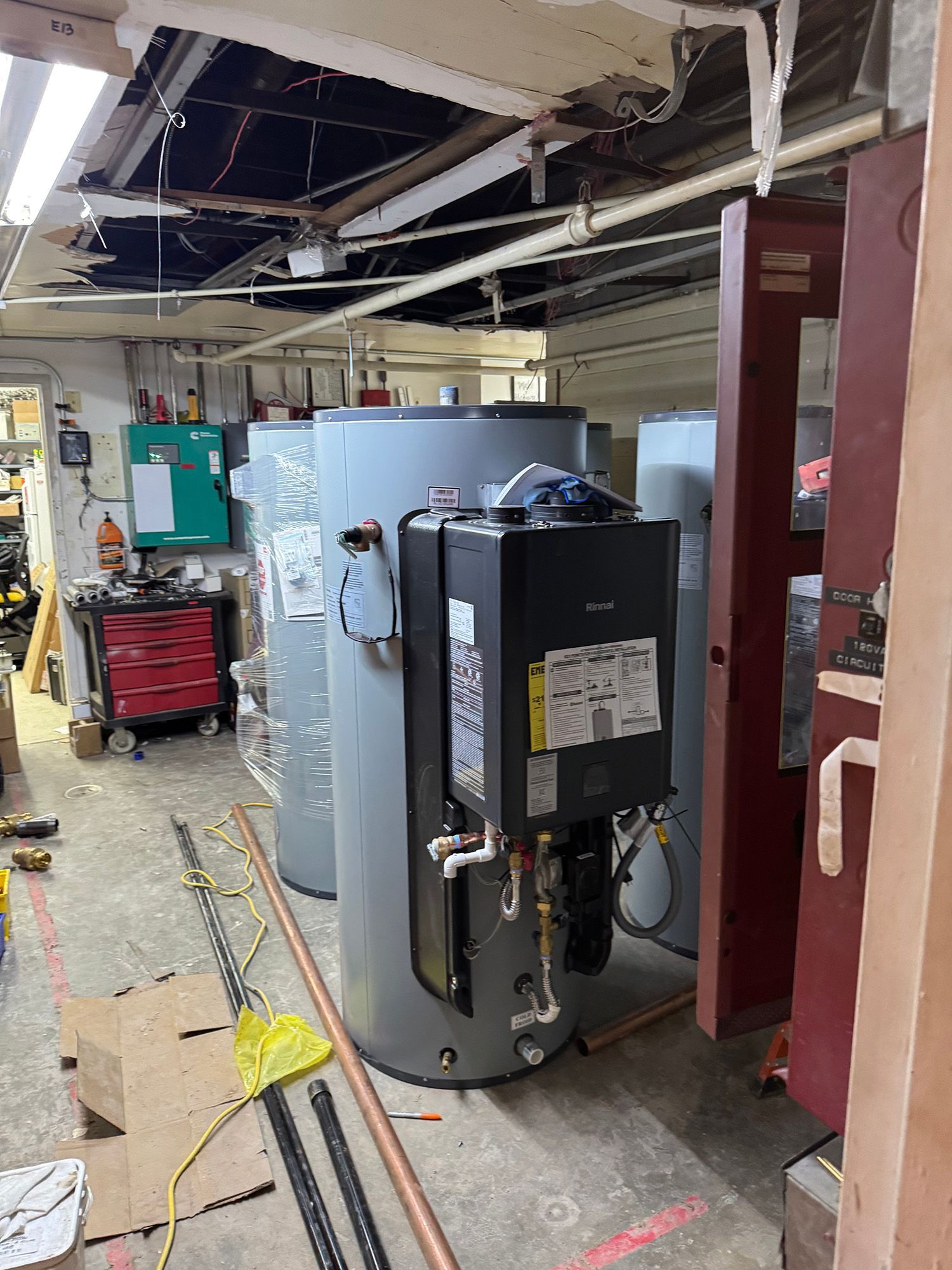 Basement with water heaters, pipes, a tool chest, and a small, green control panel.