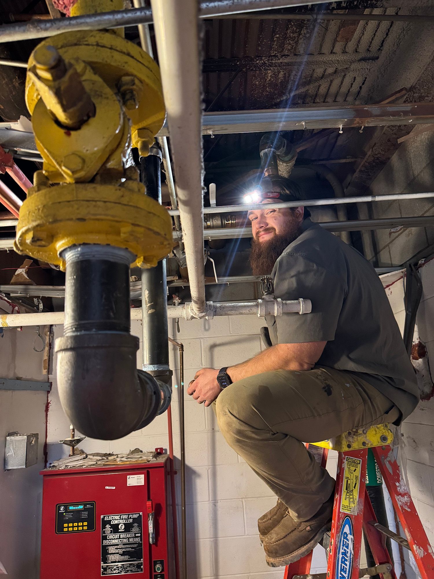 Man with headlamp on a ladder, working on pipes in a basement. Yellow and black pipe fixture. Red cabinet visible.