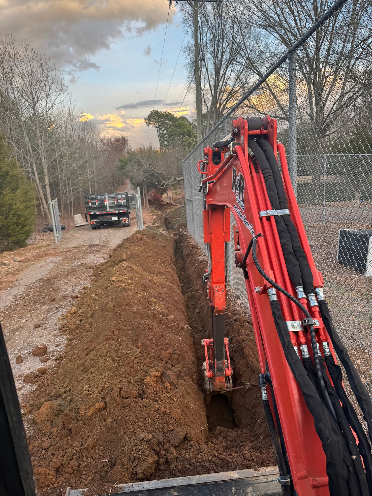 An excavator digging a trench in a dirt road, near a truck and fence, under a cloudy sky.