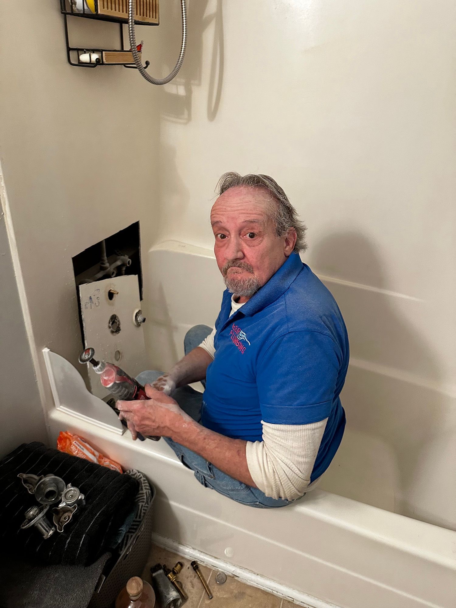 Man in blue shirt repairs shower, sitting in tub. Wall cutaway shows pipes.