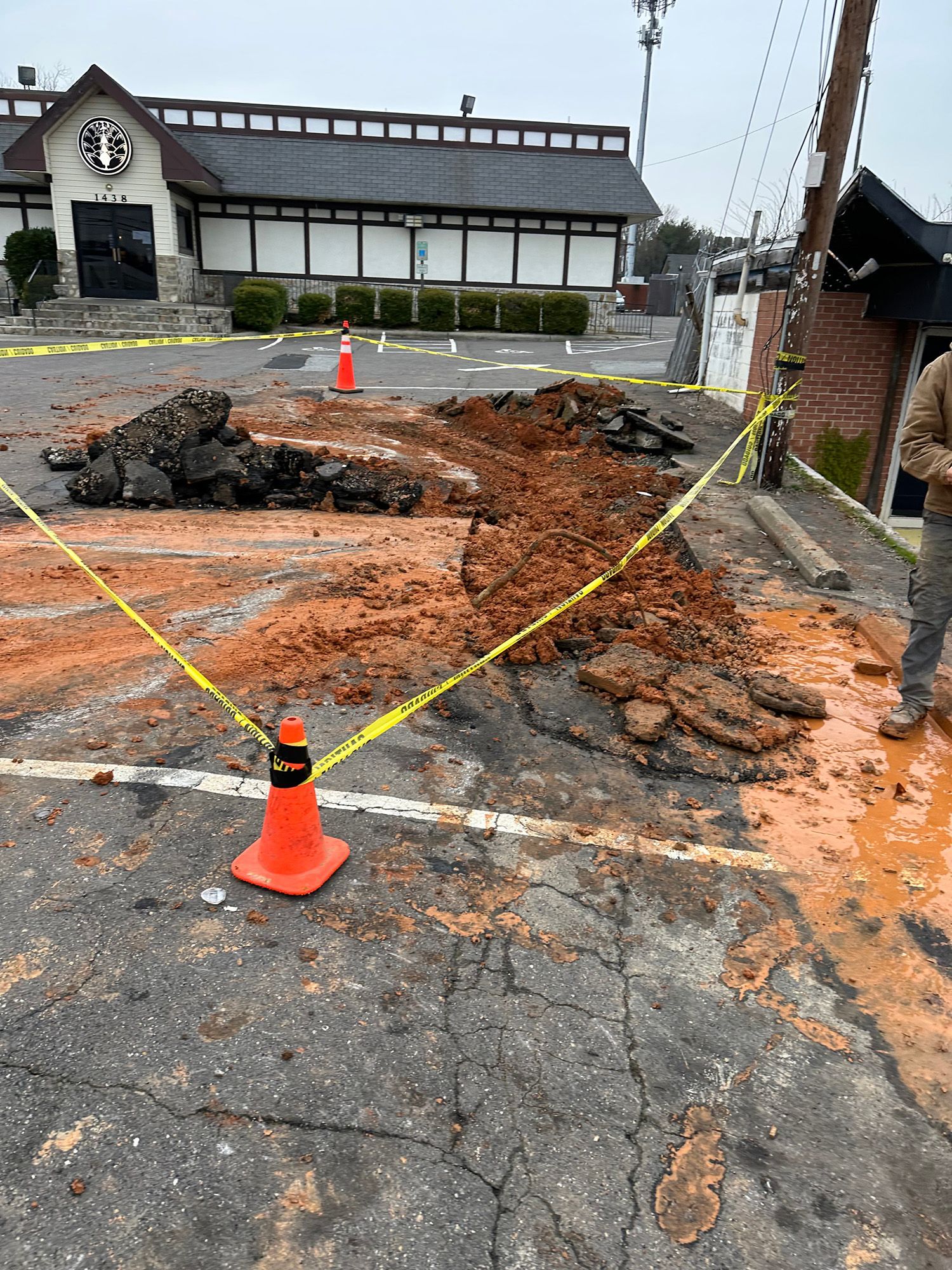 Pothole in parking lot with caution tape and debris. Restaurant building in background.