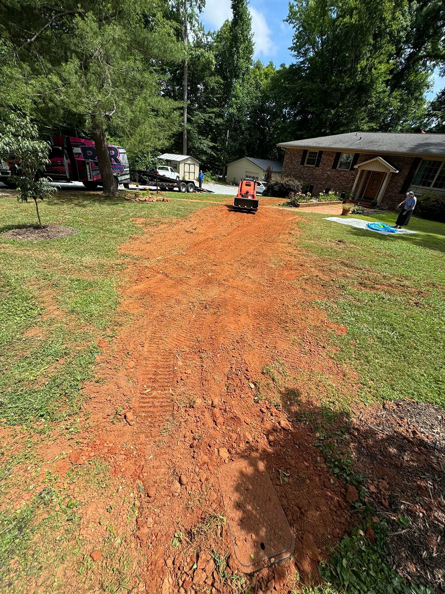 A red-dirt path in a yard with a compactor. House and trees in background.