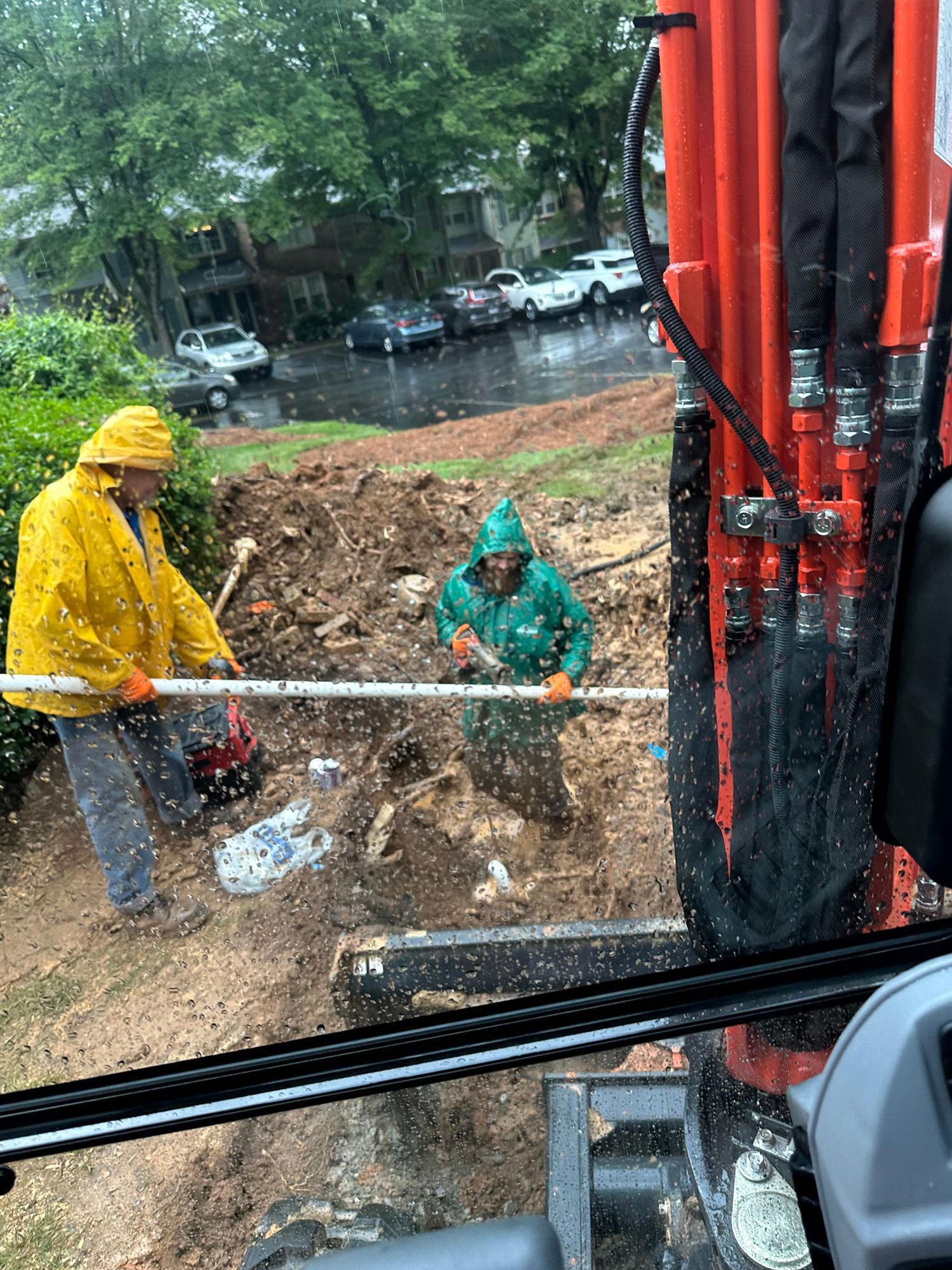Two workers in rain gear installing a pipe in a muddy ditch, orange excavator nearby.