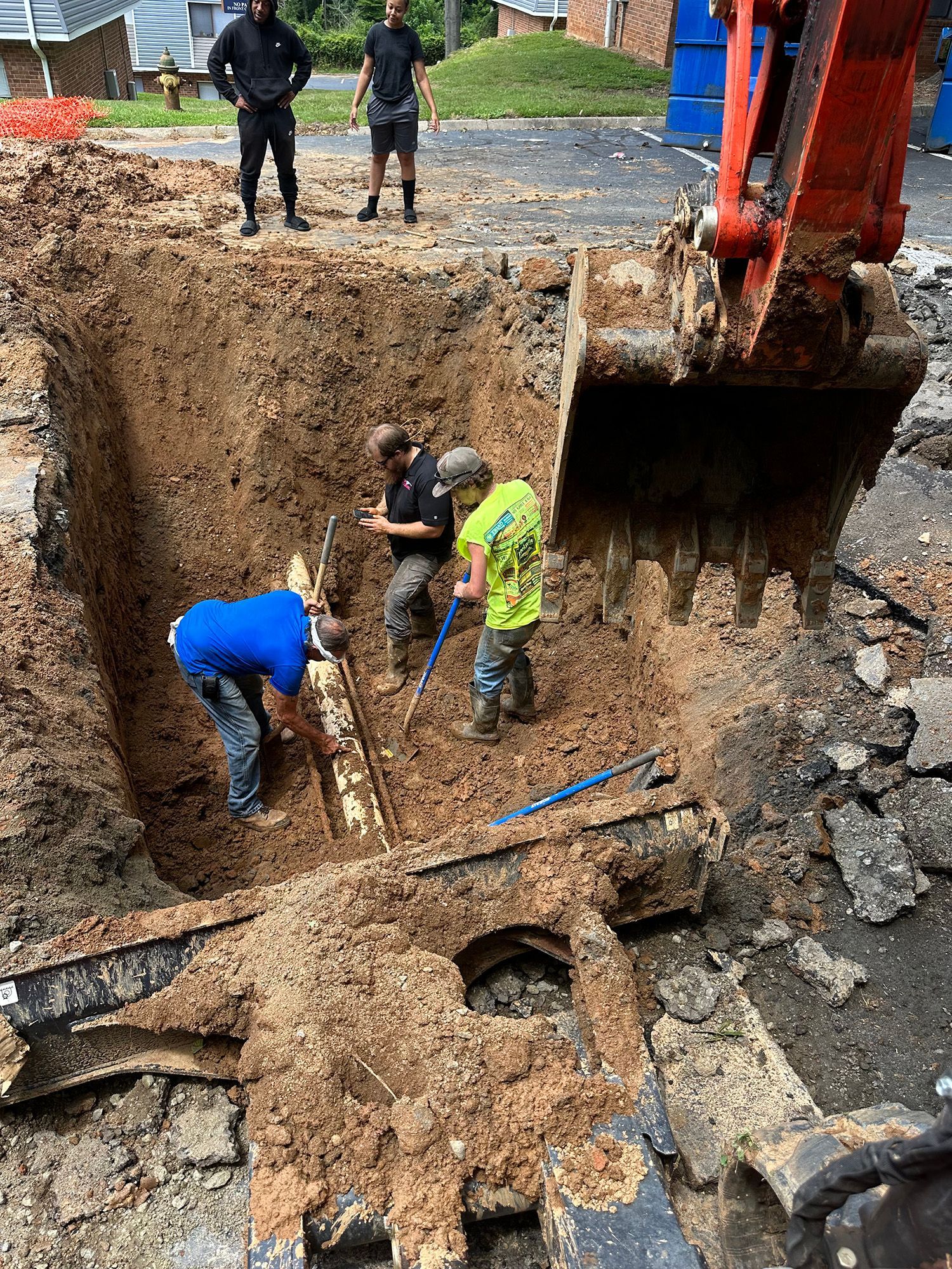 Workers repair a pipe in a trench with an excavator, while two others observe in an urban setting.