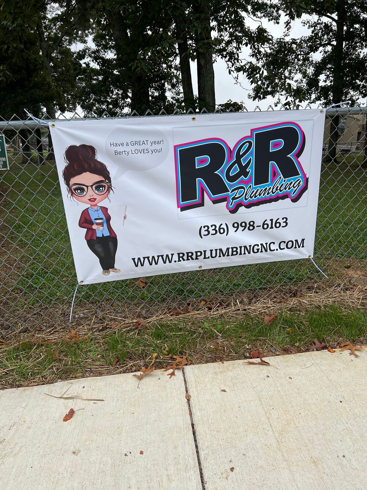Banner for R&R Plumbing, with logo and contact information, displayed on a chain-link fence.