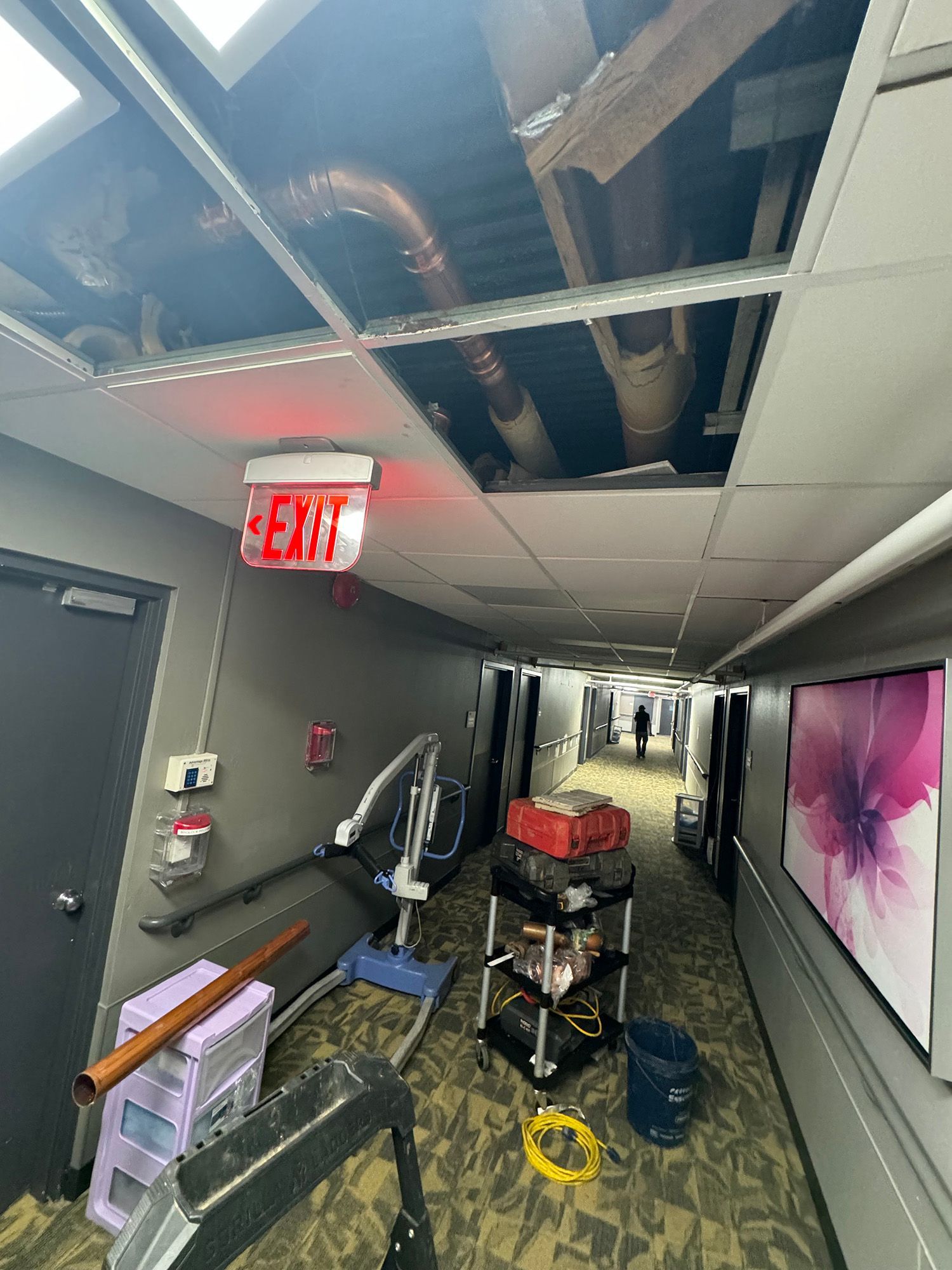 Hallway with exposed ceiling, exit sign, exercise equipment, and a person walking away.