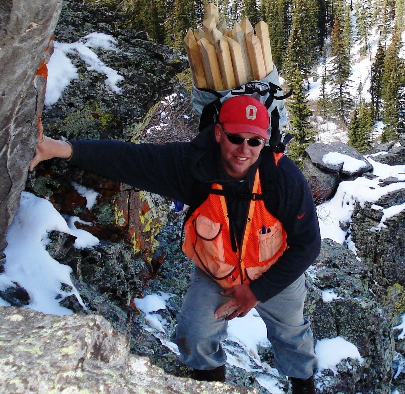Hiker in orange vest and red helmet climbing a snowy rocky slope with a backpack.