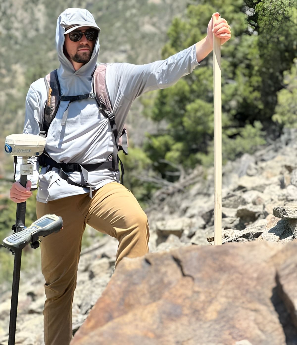 Surveyor in sun hat and sunglasses standing on rocks, holding trekking poles and GPS equipment in a mountain landscape.