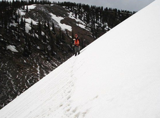 Mountaineer in red jacket climbing a steep snowy slope with pine forest in the background