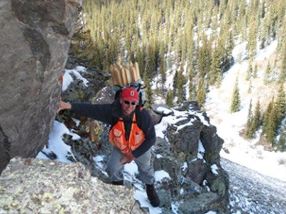Climber in orange jacket and red helmet scaling a steep snowy rock face with forest below