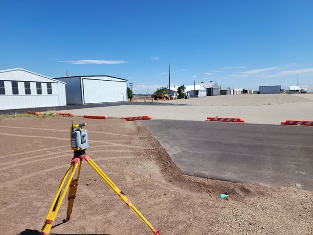 Surveying tripod with yellow legs in an open industrial lot under a clear blue sky.