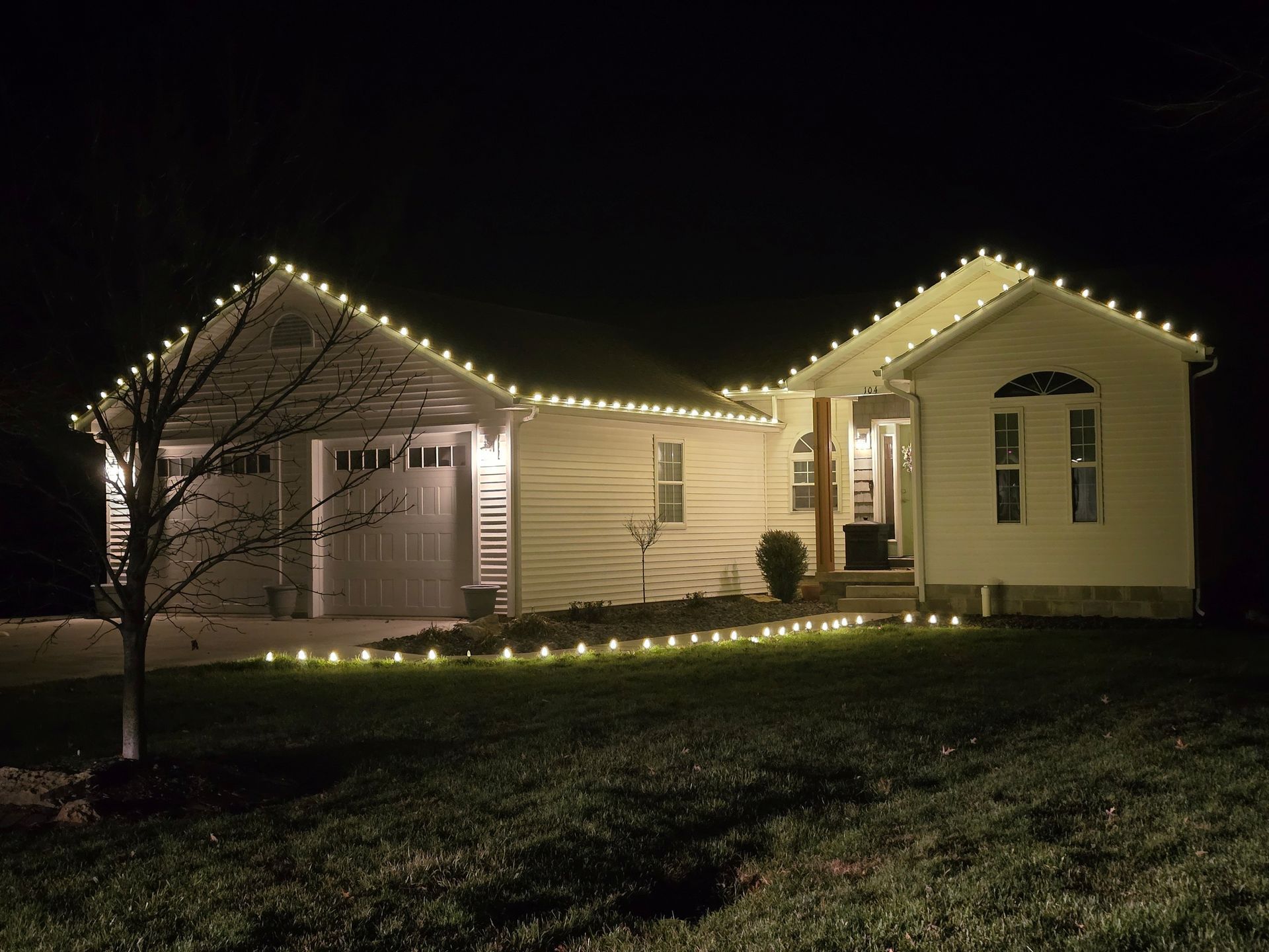 House at night illuminated with white lights along the roof and a tree.
