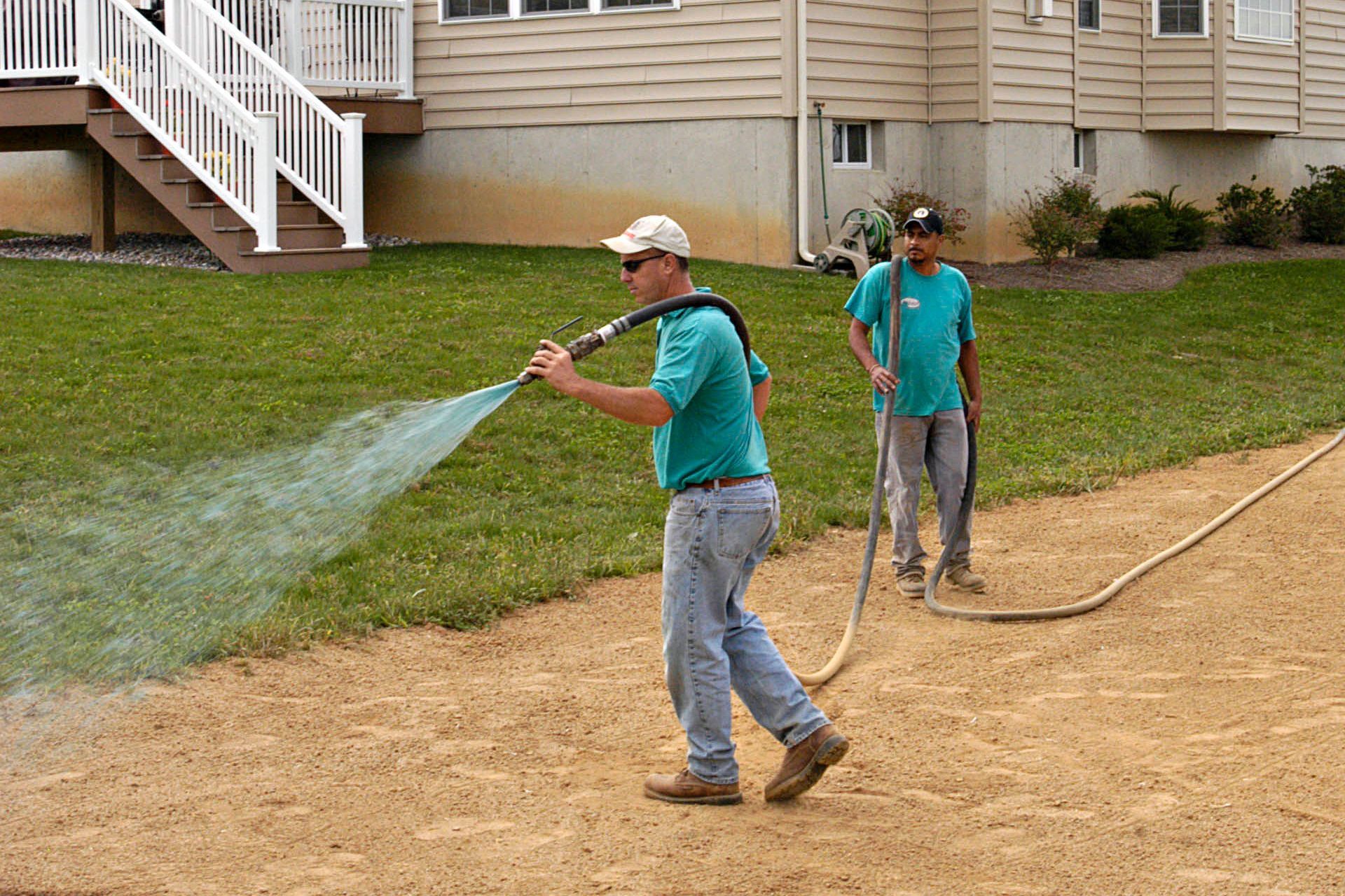 Two workers watering a lawn with hoses in front of a house. One is holding the hose and spraying water, the other is holding the hose.