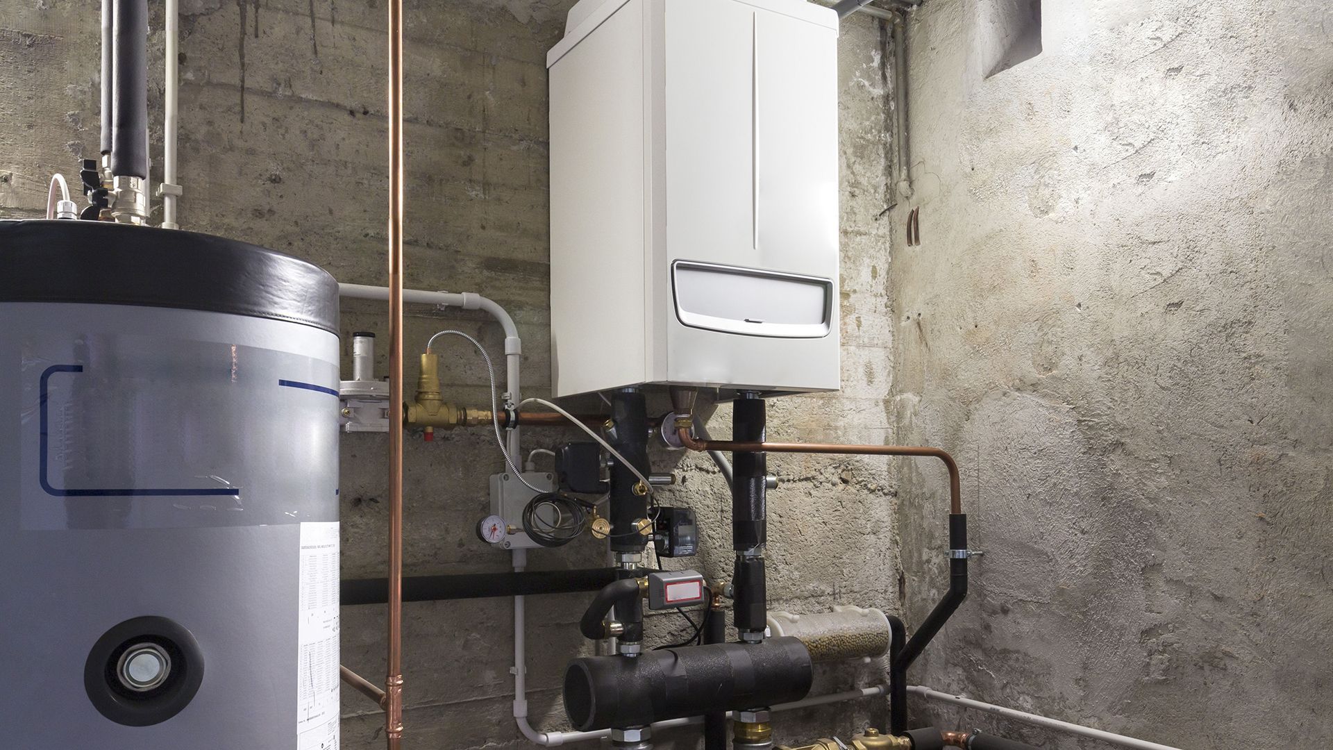 Boiler room with white wall-mounted water heater, large gray tank, pipes, and concrete walls