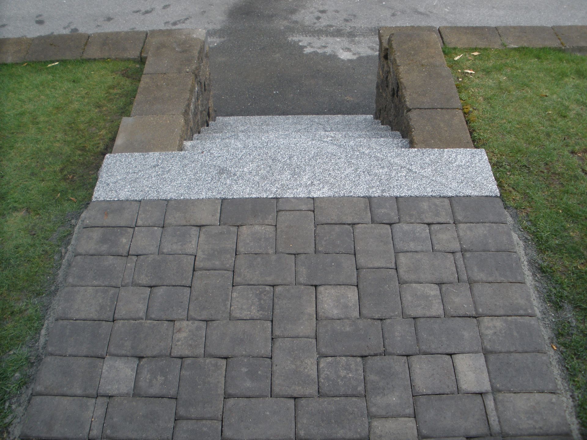 Stone steps leading down from a gray pavement, flanked by dark brick and grass.