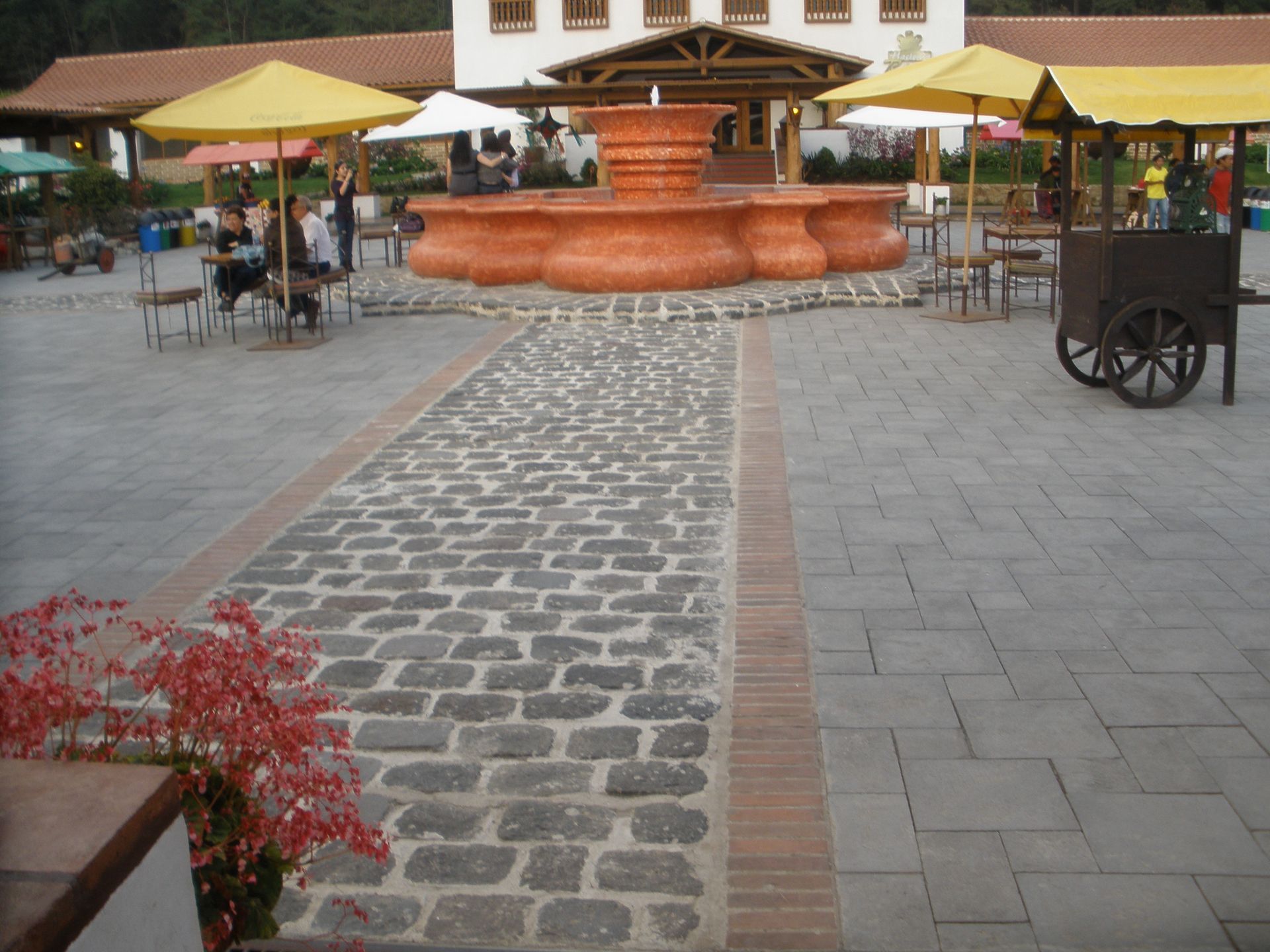 Cobblestone path leading to a large fountain in a plaza with tables, umbrellas, and a food cart.