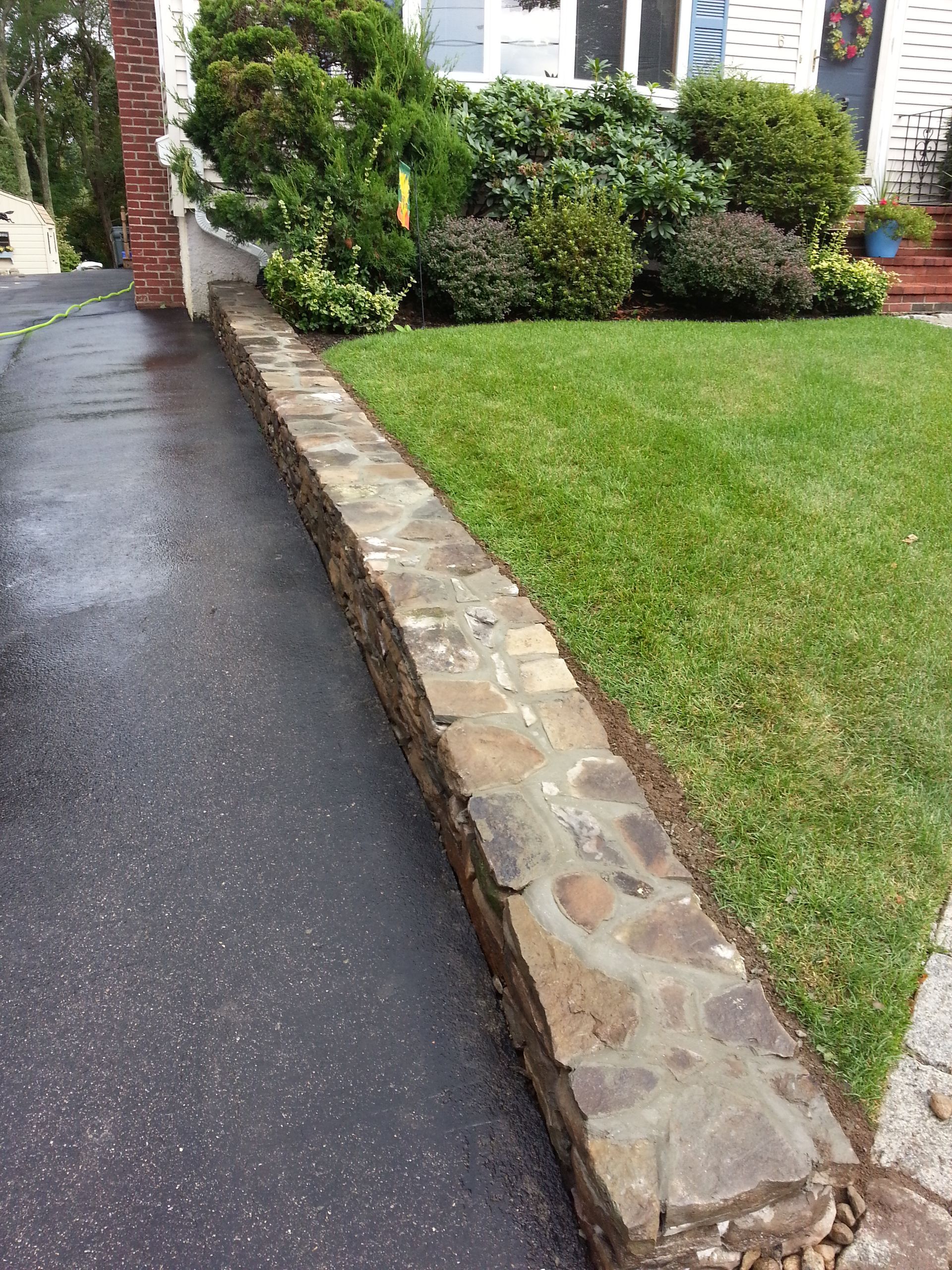 Stone retaining wall separates a driveway from a grassy lawn with shrubbery in the background.