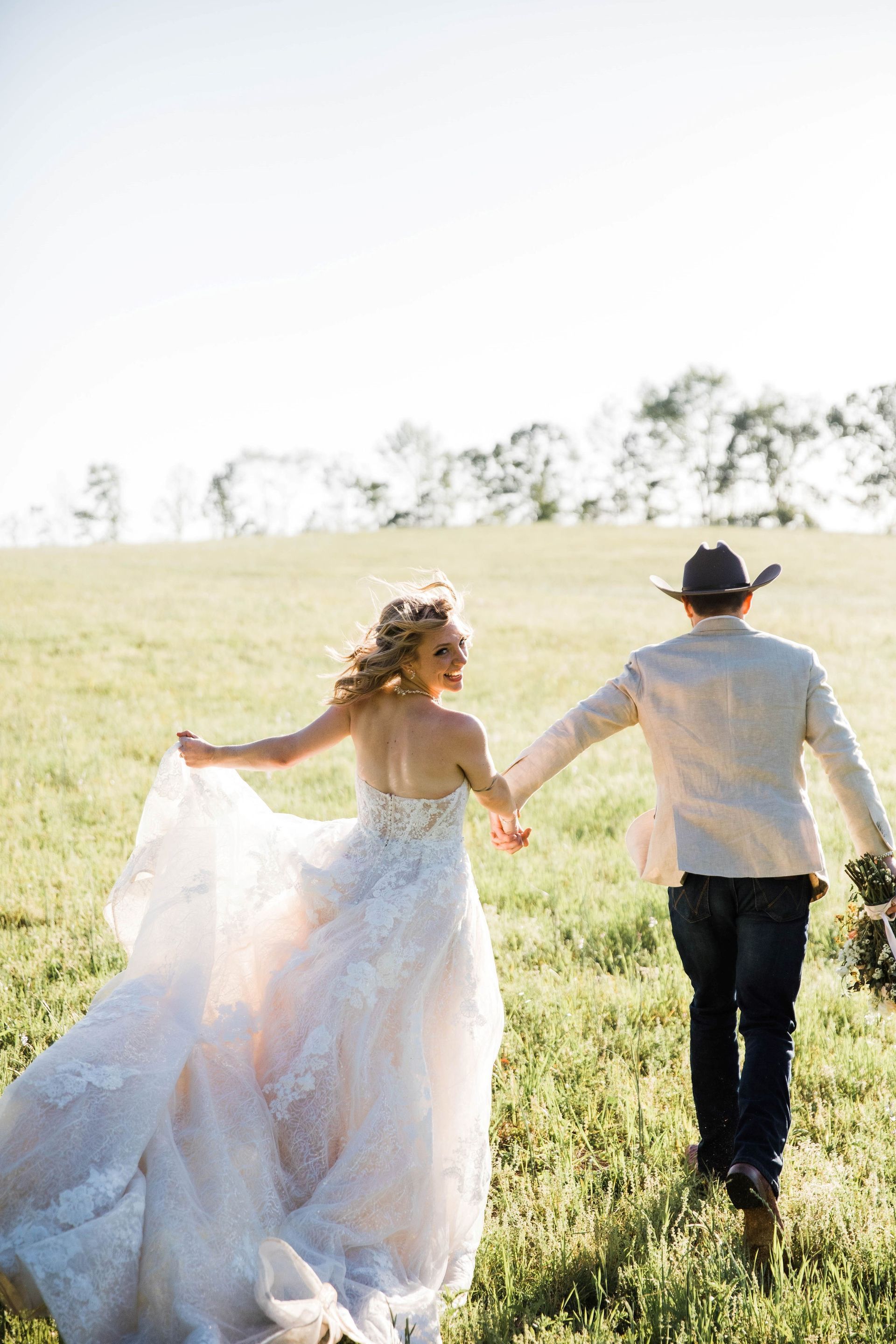 a bride and groom are running through a grassy field holding hands .