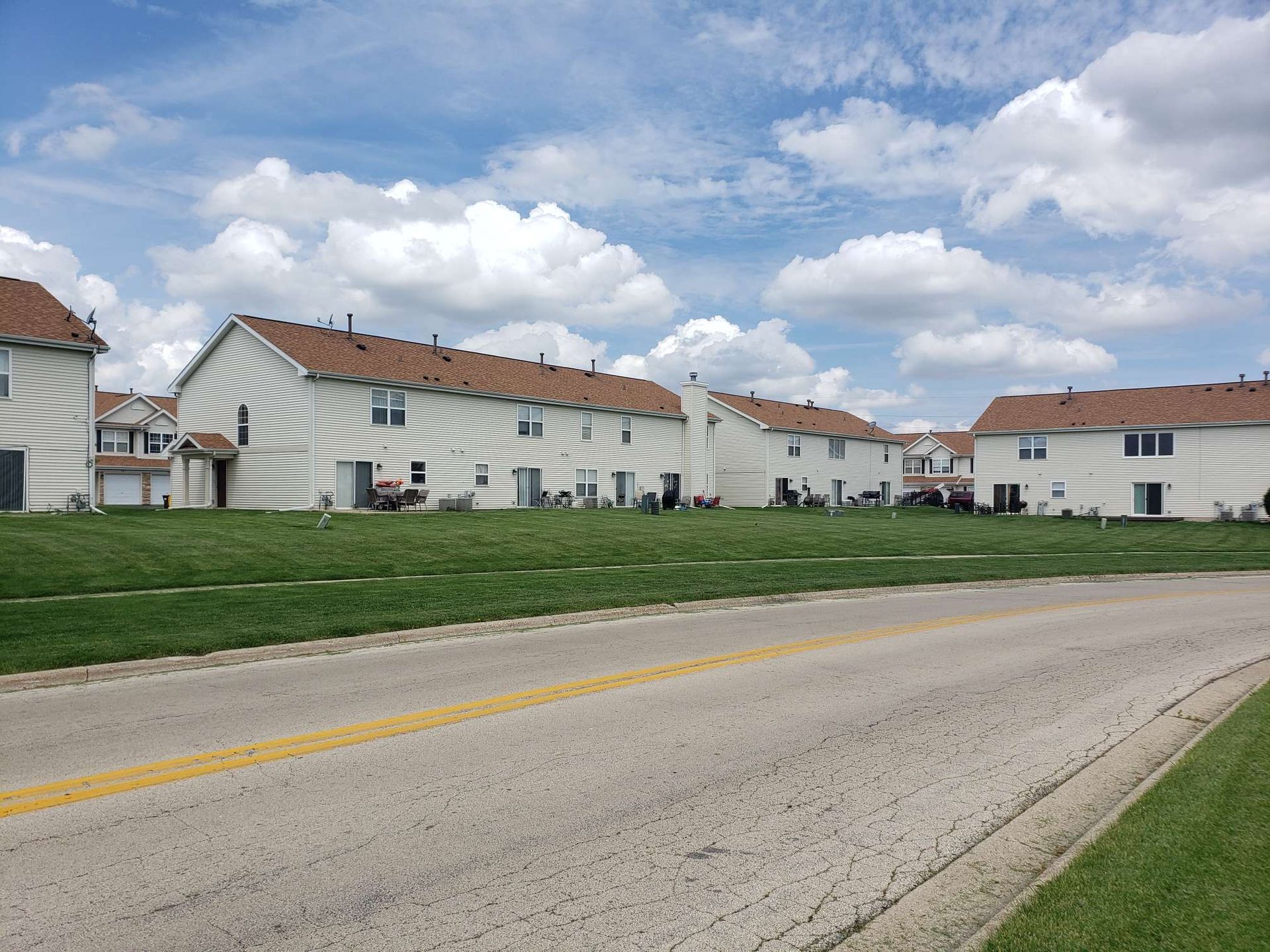 Row of two-story townhomes with brown roofs, set against a grassy lawn and a road, under a cloudy sky.