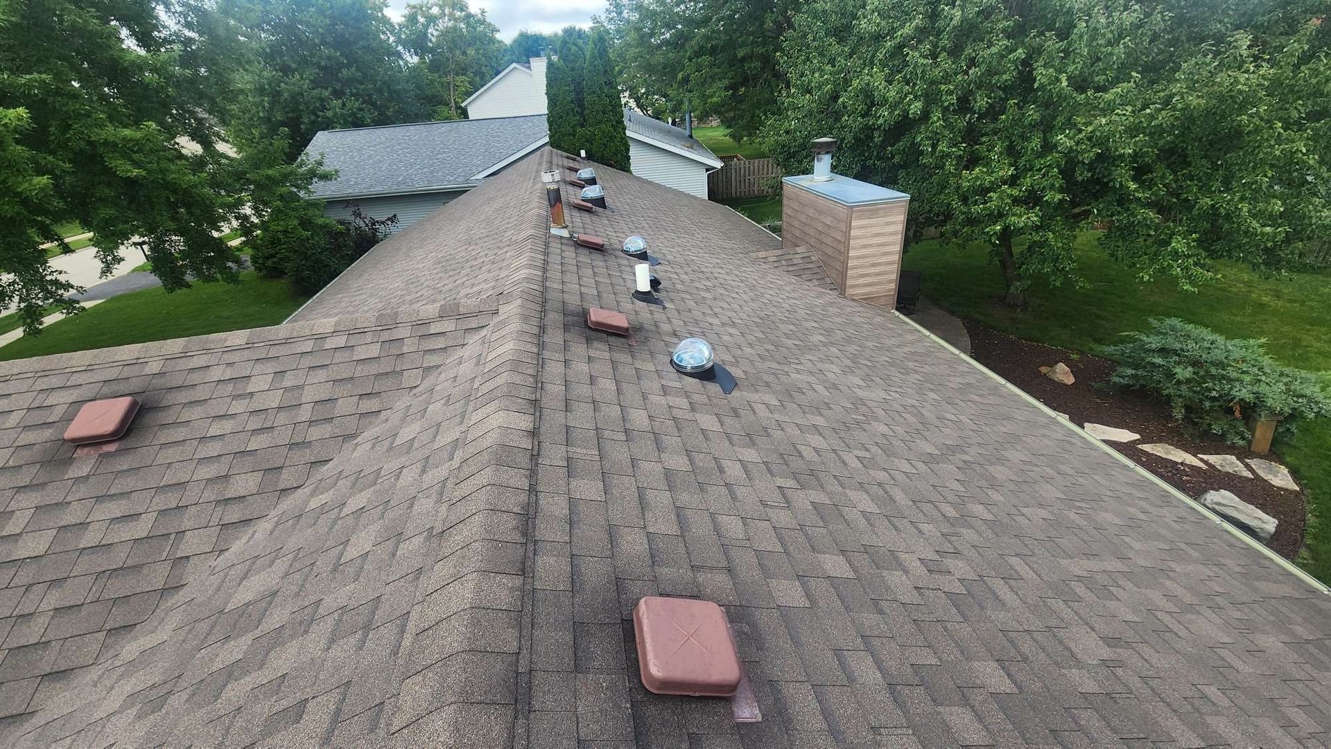 Overhead view of a brown shingle roof with vents, a brick chimney, and trees in the background.