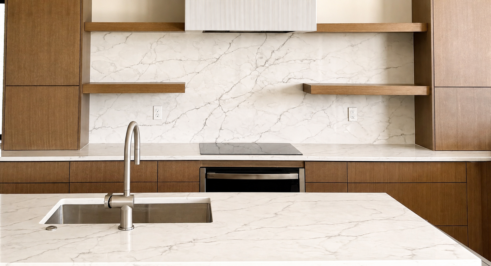 Modern kitchen with white marble island, wood cabinetry, built-in appliances, and stainless-steel sink.