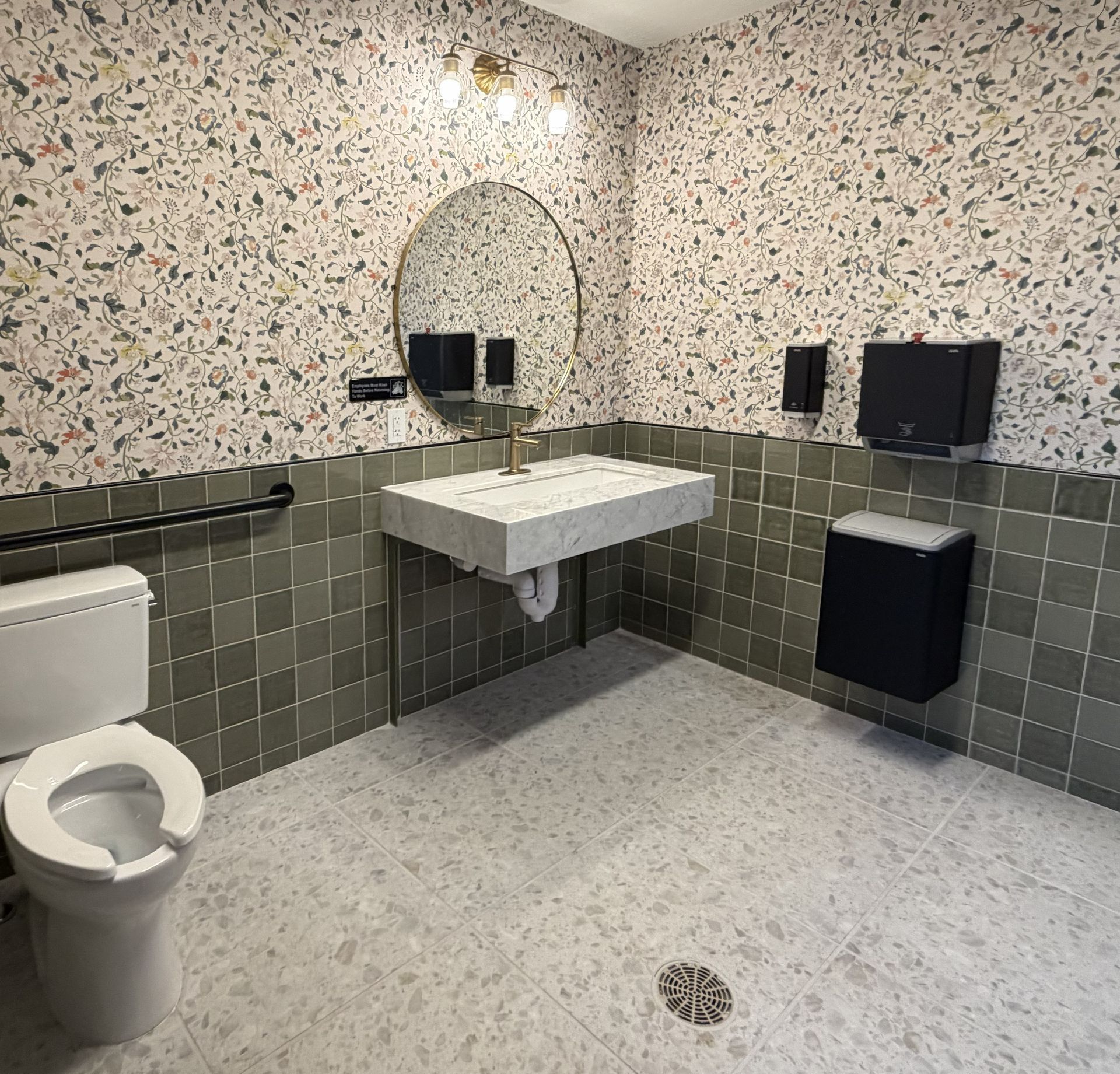Public restroom with green tile, floral wallpaper, sink, mirror, toilet, and hand dryer.