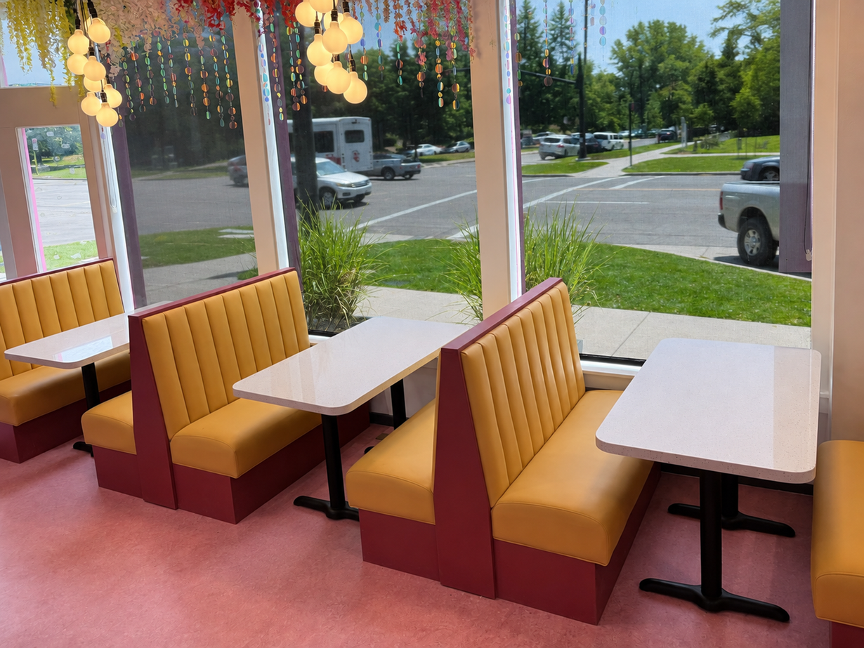 Booth seating by large windows in a bright diner, with white tables and yellow-orange benches.
