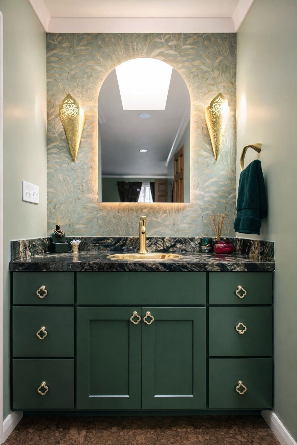 Dark green bathroom vanity with marble countertop, brass fixtures, arched mirror, and wall sconces