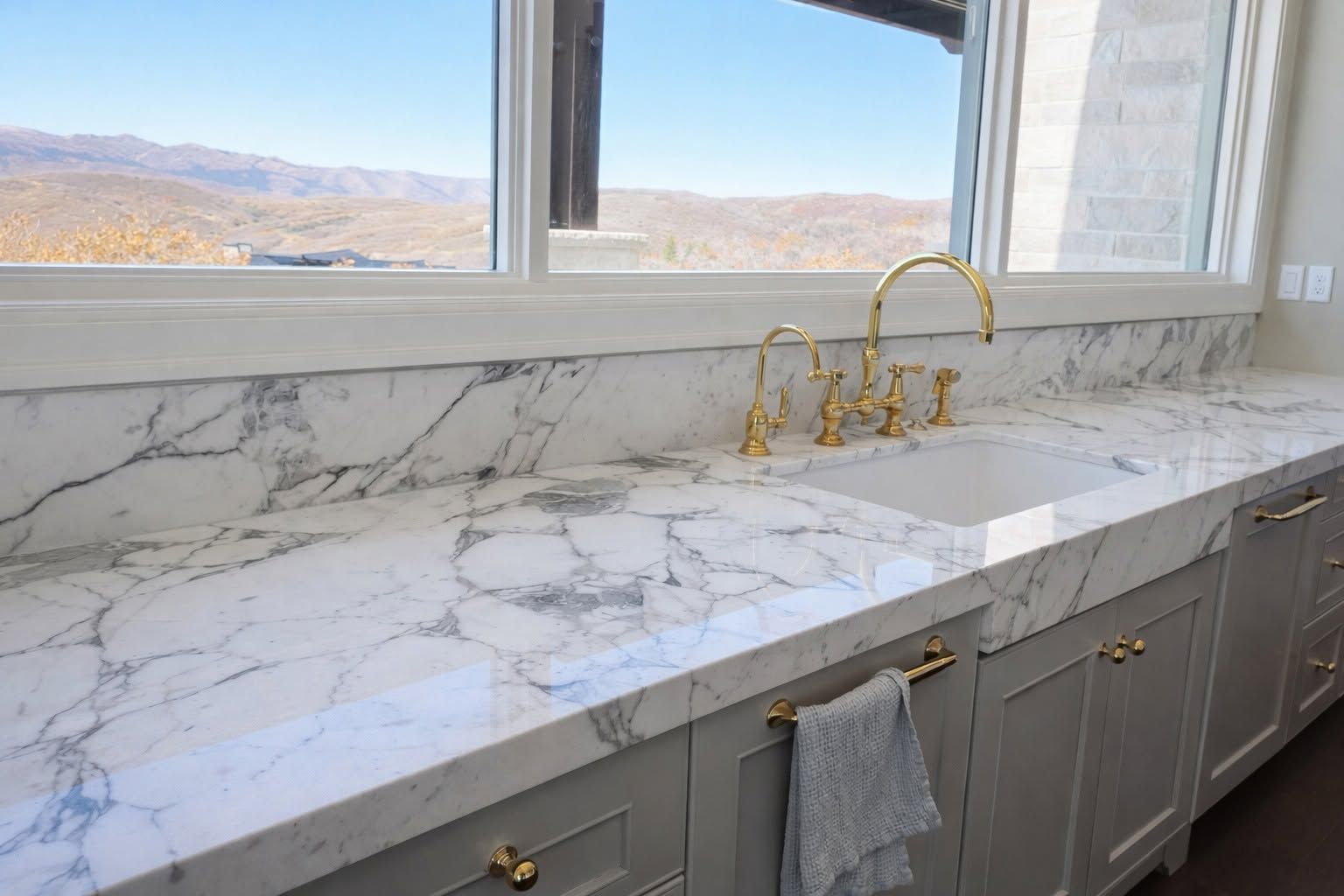Marble bathroom vanity with gold faucets beneath a window overlooking desert mountains