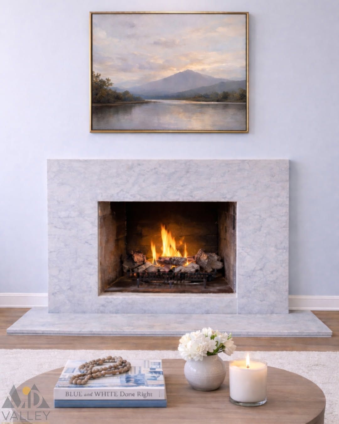 Modern living room with a lit fireplace, landscape painting, and coffee table with books, flowers, and a candle