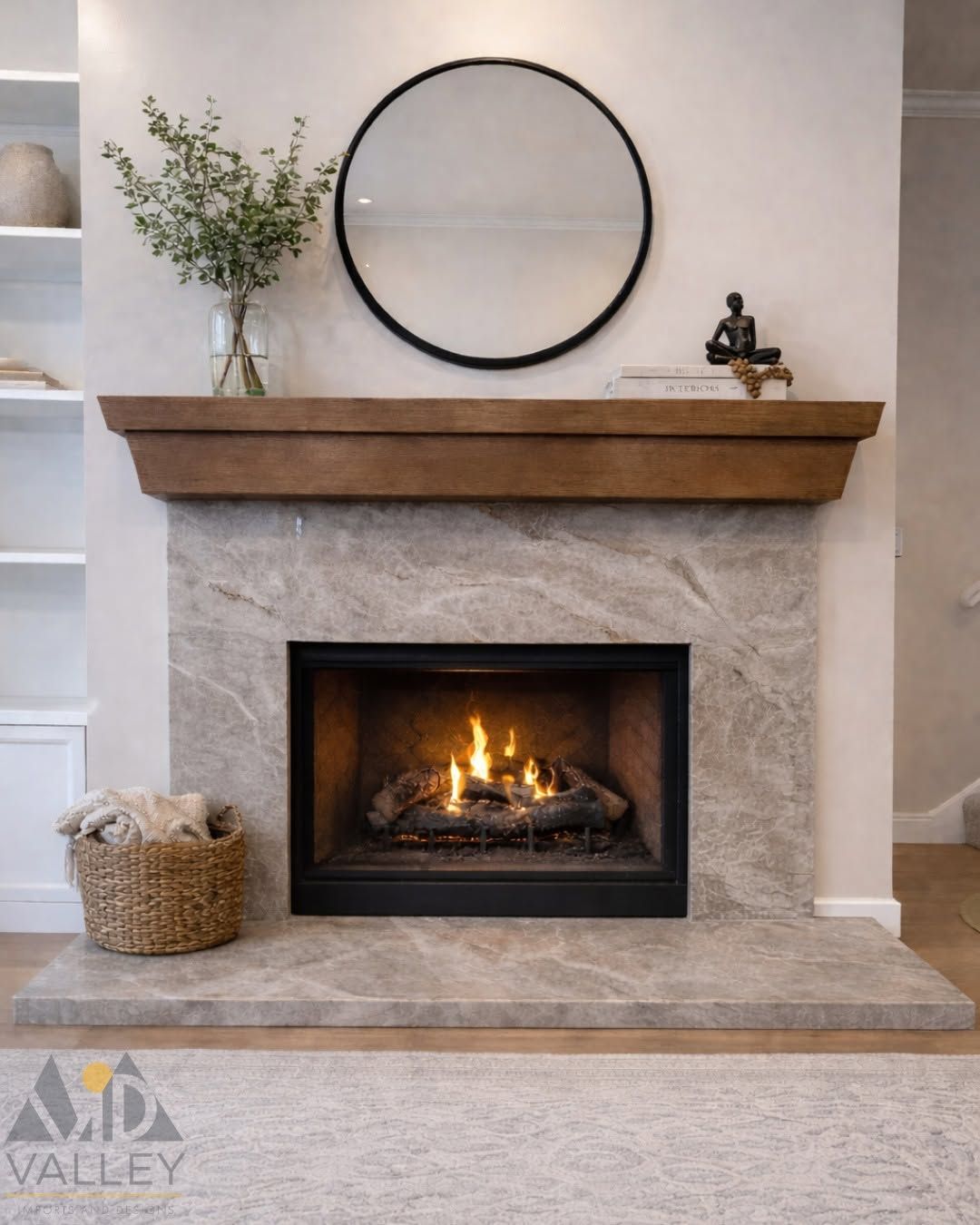 Stone fireplace with a lit fire, wood mantel, round mirror, vase, and basket in a living room.