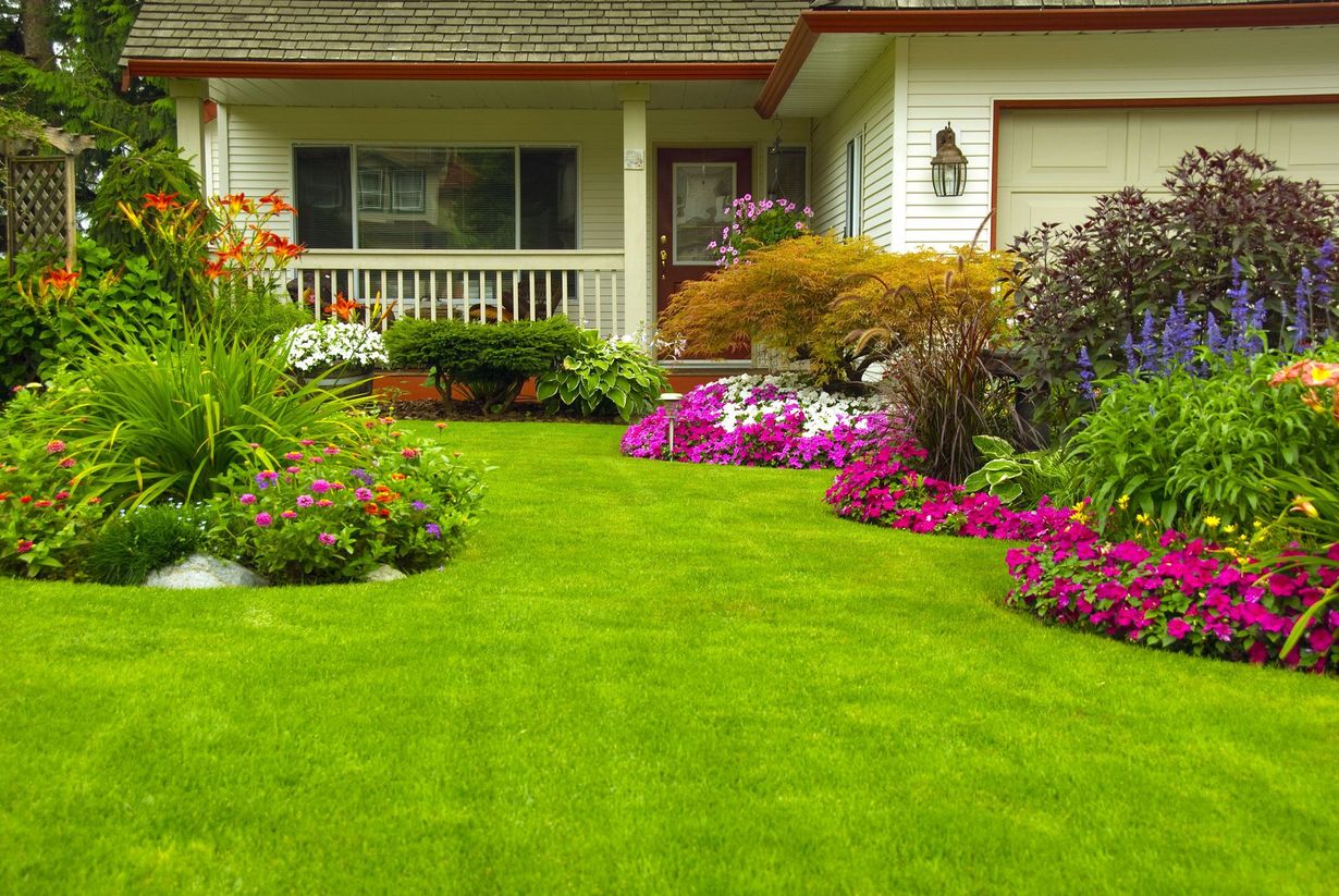 Lush green lawn with colorful flower beds in front of a white house with a porch and garage.