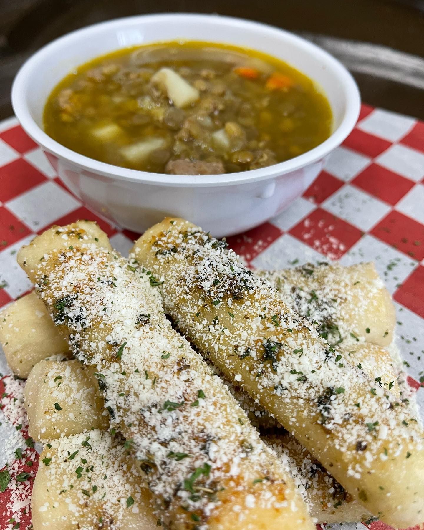 A bowl of soup and garlic sticks on a checkered table cloth.