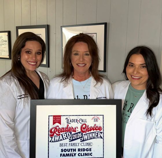 Learn More About Family Practice Three women in white coats holding a South Ridge Family Clinic award plaque, smiling indoors