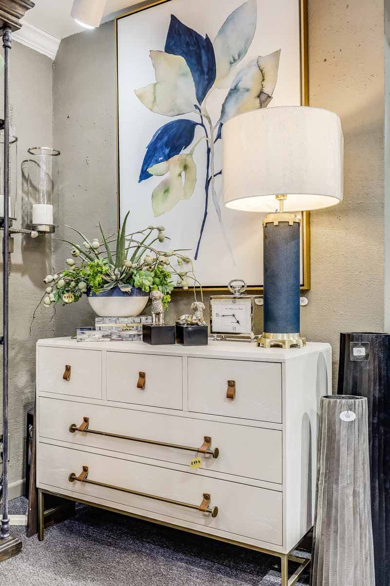 Elegant dresser with floral wall art, blue lamp, and potted greenery in a bright room.