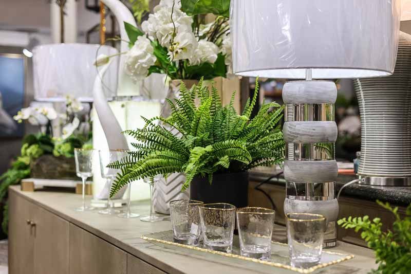 Glass table display with white lamps, green potted plants, and small glass cups in a bright indoor setting
