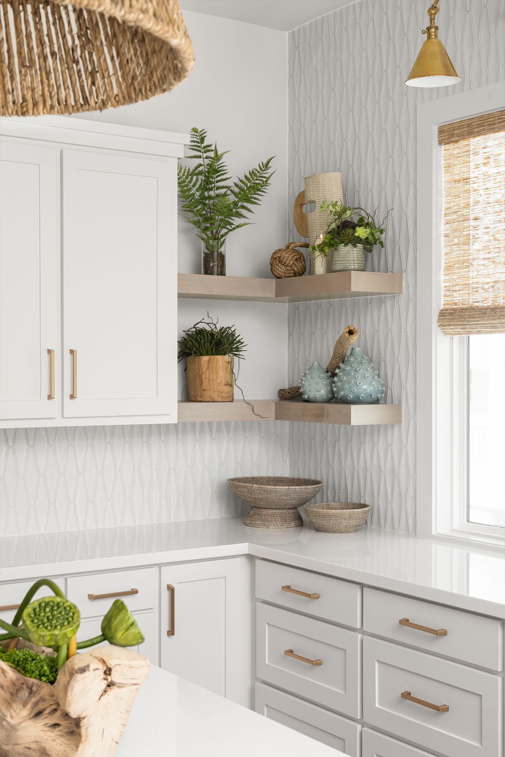 Bright white kitchen corner with gold accents, open shelves, potted plants, woven baskets, and a window with shades