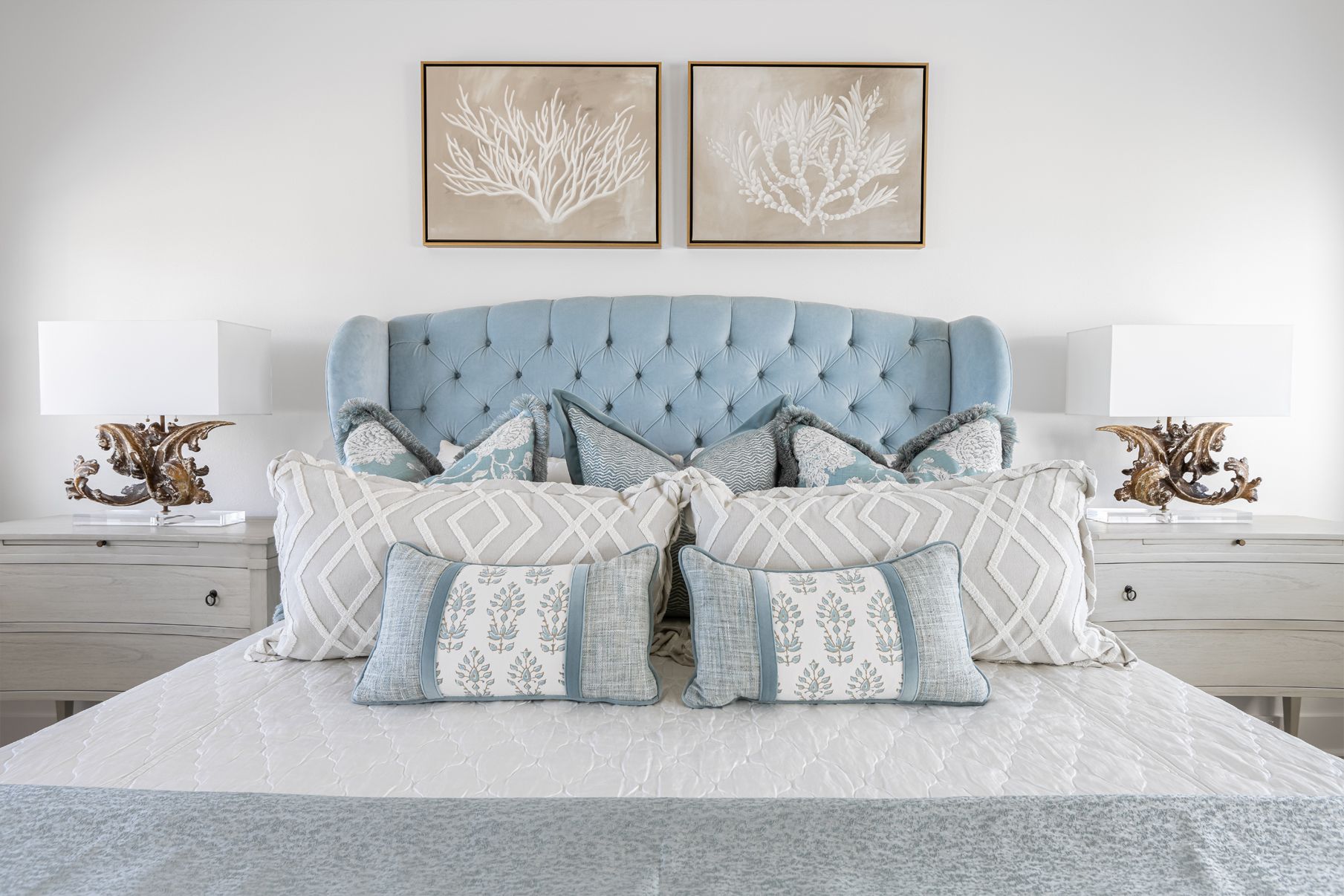 Light blue tufted bed with patterned pillows and white lamps in a bright, symmetrical bedroom
