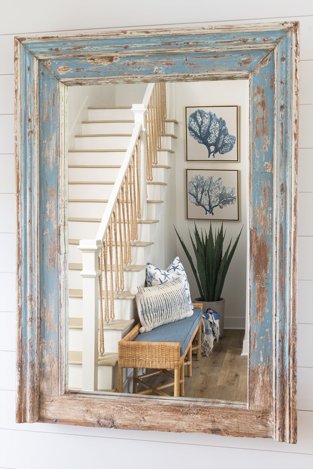 Rustic blue mirror reflecting a bright entryway with staircase, bench, framed art, and potted plant