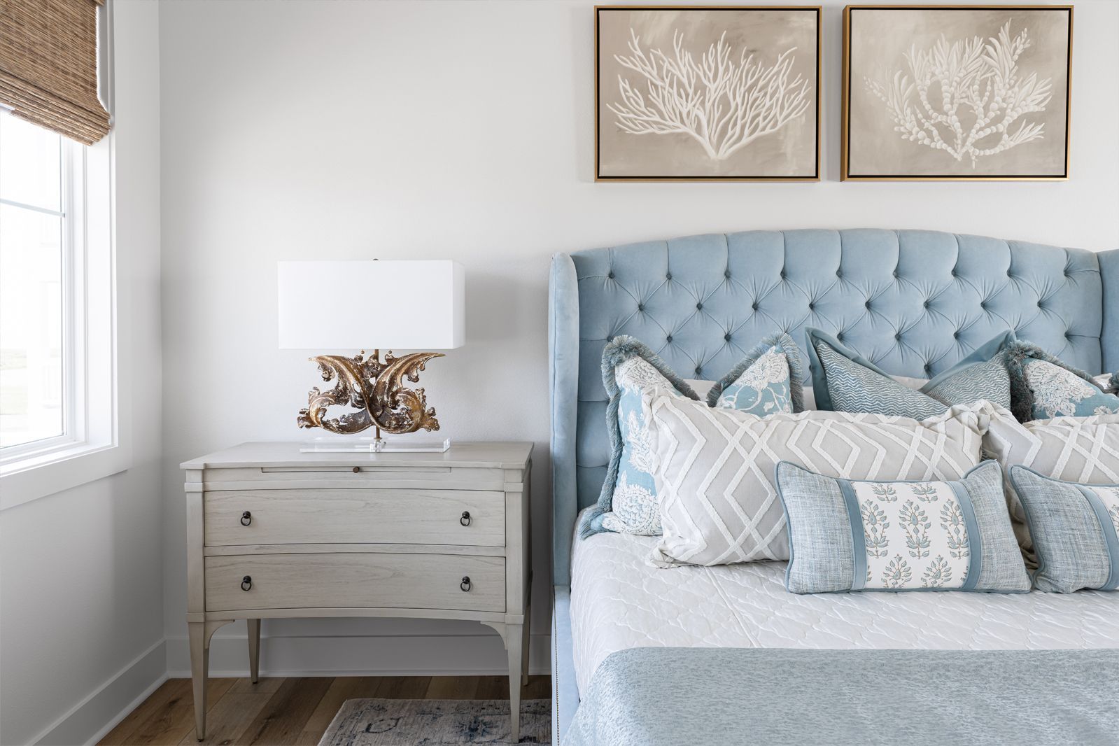 Bright bedroom with a tufted blue headboard, white nightstand, and beige wall art by a window