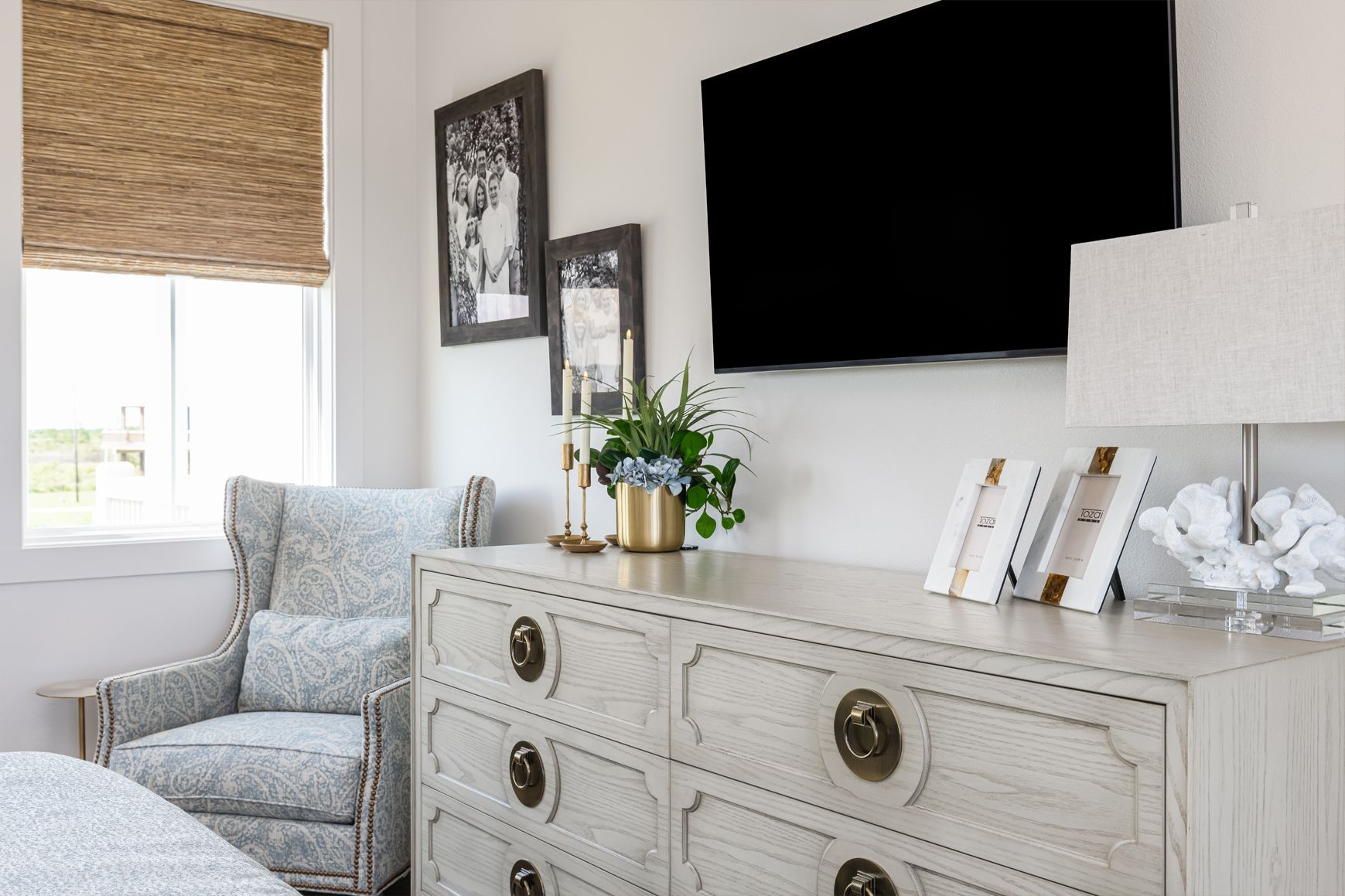 Bright living room with a gray armchair, white dresser, wall-mounted TV, and window with woven shade