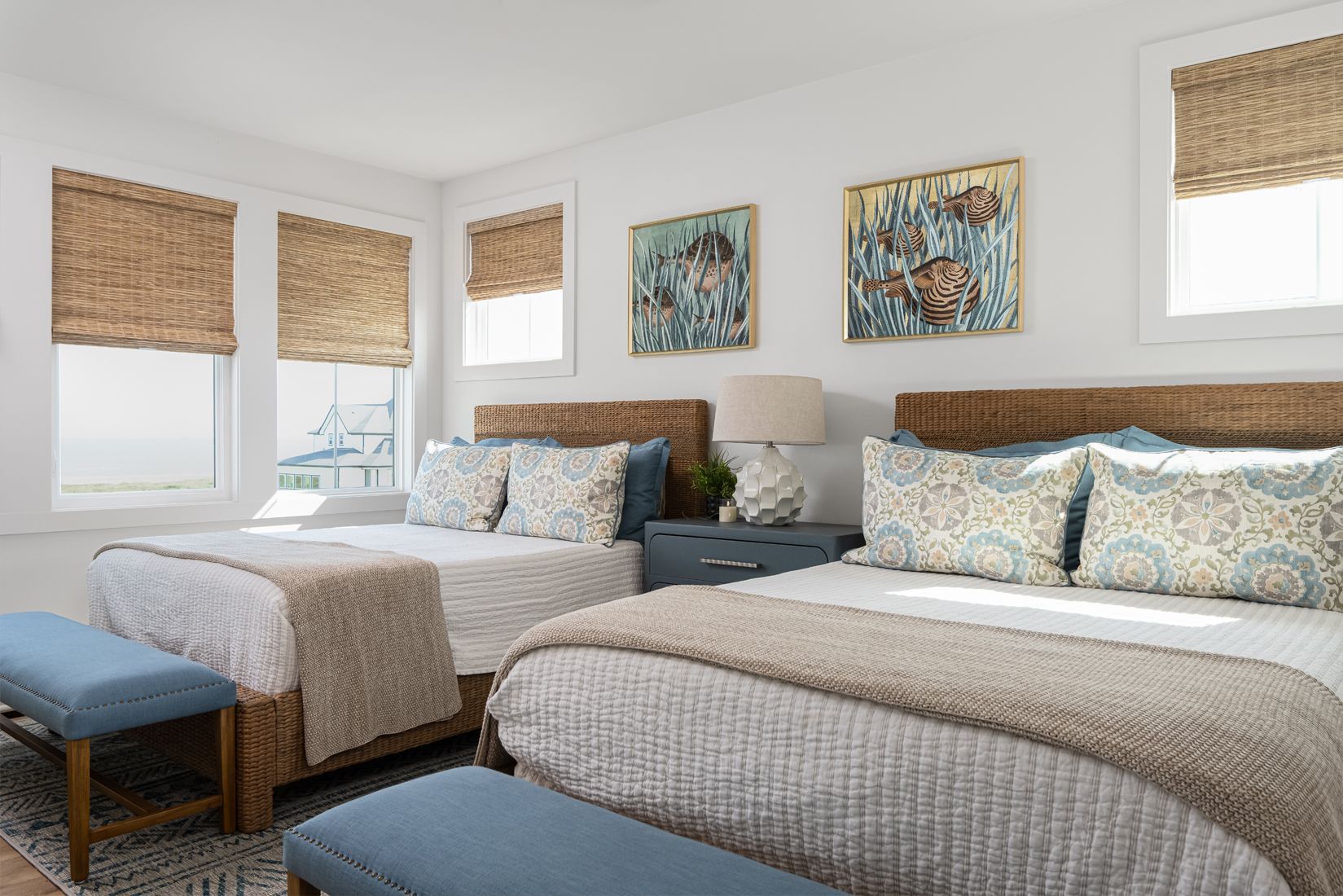 Bright bedroom with two beds, blue bench, and woven shades over large windows.
