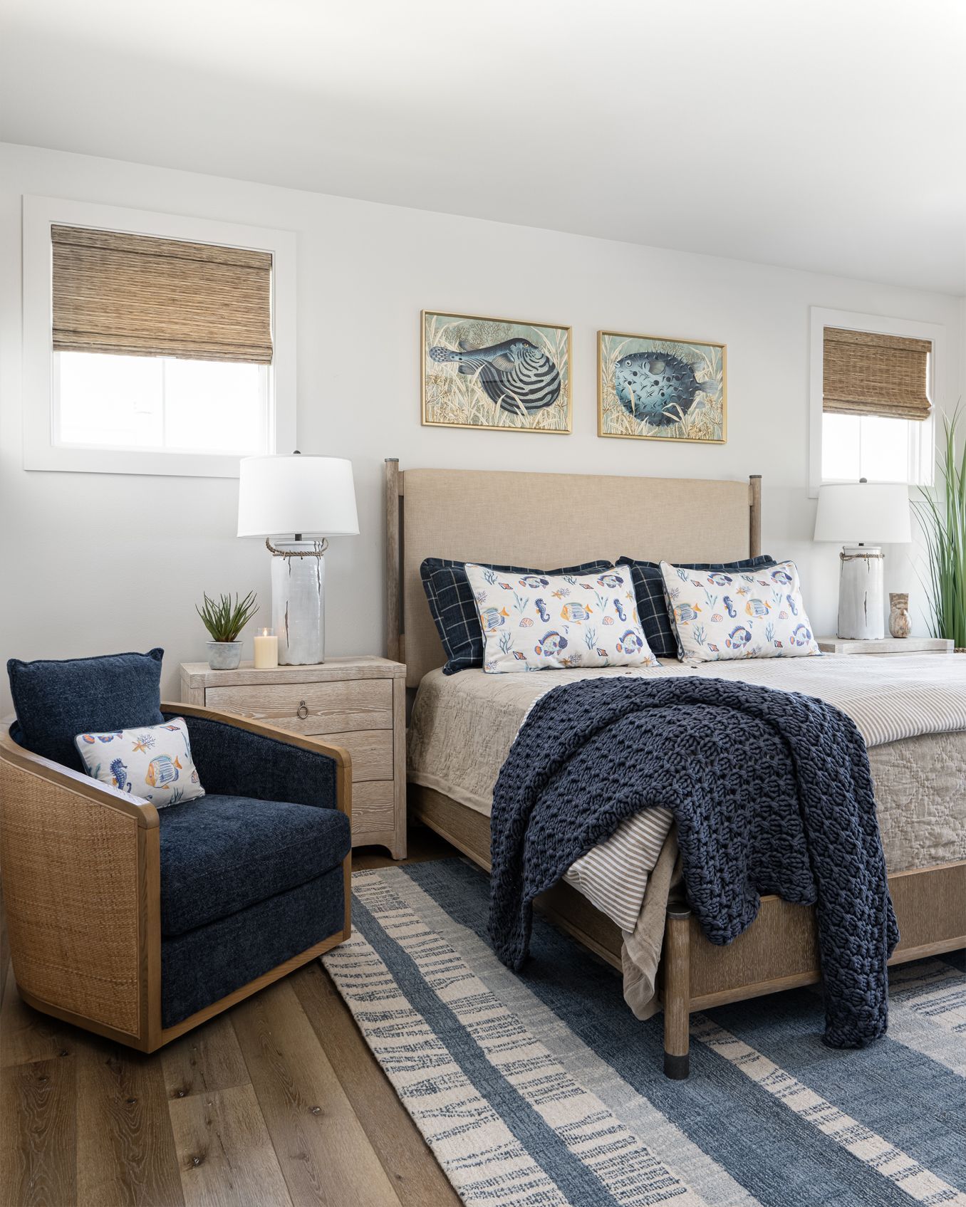Coastal-style bedroom with beige bed, navy accents, wicker chair, patterned rug, and ocean art on white walls