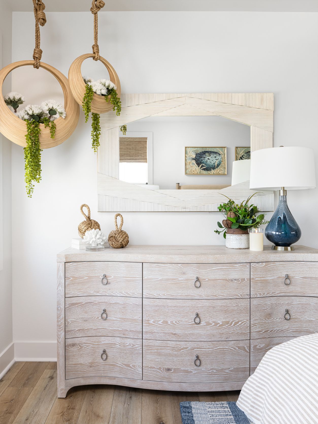 Light-filled bedroom with white dresser, round mirrors, hanging plants, and blue lamp